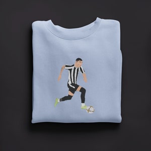 Puede incluir: Una sudadera de color azul claro con un gr&aacute;fico de un jugador de f&uacute;tbol con una camiseta a rayas blancas y negras regateando un bal&oacute;n de f&uacute;tbol.