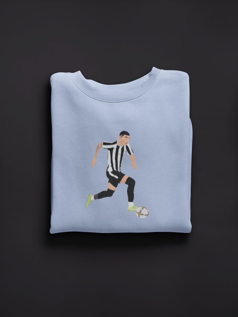 Puede incluir: Una sudadera de color azul claro con un gr&aacute;fico de un jugador de f&uacute;tbol con una camiseta a rayas blancas y negras regateando un bal&oacute;n de f&uacute;tbol.