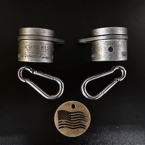 May include: An assortment of metal accessories, including two cylindrical pieces with the text "CONGRESS" and "2026", two silver carabiner clips, and a round tag with an engraved American flag. The items are arranged on a dark surface.