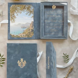 Peut inclure: Un coffret d'invitation de mariage en velours bleu avec une photo de paysage côtier sur le panneau gauche et une invitation sous verre avec texte doré à droite. La couverture extérieure présente un blason doré et le texte THE WEDDING.