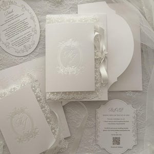 May include: A white wedding invitation suite featuring embossed floral oval frames with the initials MJ, intricate lace borders, and satin ribbon accents. Includes a circular details card, a rectangular RSVP card with a QR code, and envelopes.