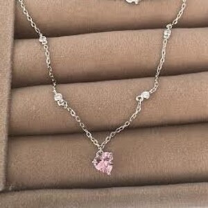 May include: A silver chain necklace with a small pink heart pendant. The necklace is displayed on a brown velvet background.