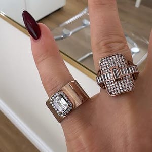 May include: Close-up of two ornate rings. One ring features a rectangular, clear gemstone surrounded by smaller stones, set on a gold band. The other ring has a square design with multiple clear stones. The rings are on a finger with a burgundy nail.