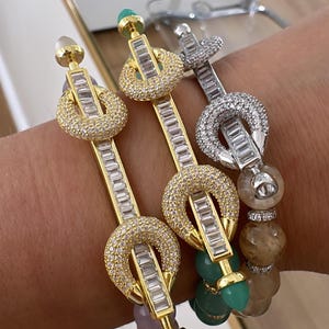 Può includere: Tre braccialetti con finitura oro o argento, caratterizzati da un design unico. Ogni braccialetto ha una barra centrale con pietre rettangolari e un elemento circolare con pietre più piccole. I braccialetti hanno perline colorate sul fondo.