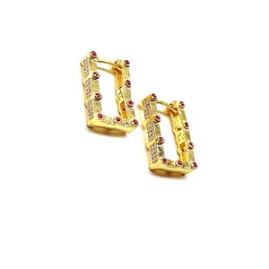 以下が含まれることがあります： 正方形のデザインのゴールドのフープピアス。ピアスは、小さなクリアなキュービックジルコニアと赤い宝石で飾られています。