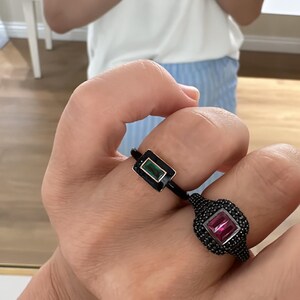 Puede incluir: Dos anillos con engastes negros y piedras preciosas de colores. El anillo de la izquierda tiene una piedra rectangular verde, mientras que el anillo de la derecha tiene una piedra rectangular rosa.
