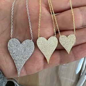 May include: Three silver and gold necklaces with heart-shaped pendants. The pendants are covered in small, clear stones. The necklaces are on a hand.
