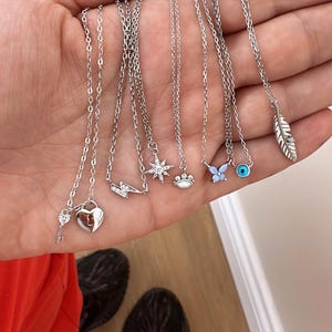May include: A collection of silver necklaces with various charms, including a heart with a key, a lightning bolt, a star, a paw print, a butterfly, an evil eye, and a feather.