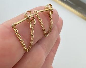 Puede incluir: Un par de pendientes de botón dorados con un diseño de barra horizontal conectado a una delicada cadena colgante que vuelve al poste. Los pendientes tienen un acabado metálico pulido e incluyen cierres de mariposa estándar.