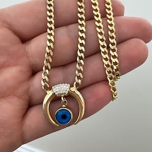 May include: A gold chain necklace with a blue evil eye charm pendant. The charm is surrounded by small clear stones and has a crescent moon shape.