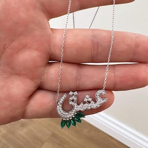 May include: A silver necklace with a pendant spelling out a word in Arabic script. The pendant is encrusted with clear crystals and has a cluster of green emerald-cut stones at the bottom. The necklace is displayed against a neutral background.