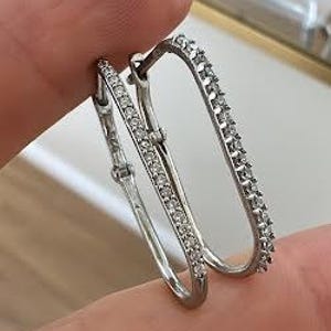 May include: Silver-toned hoop earrings with a rectangular shape. One earring is adorned with small, clear gemstones along the outer edge, while the other has a single row of gemstones. The earrings are held between fingers.