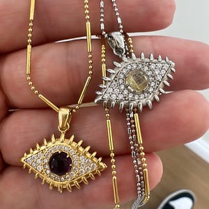 May include: Two eye-shaped pendant necklaces. One is gold-toned with a purple center stone and clear accent stones. The other is silver-toned with a yellow center stone and clear accent stones. Both have delicate chains.