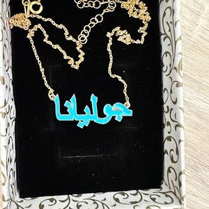 May include: A gold chain necklace with a blue nameplate pendant that reads "جوانا". The necklace is in a white box with a floral design.