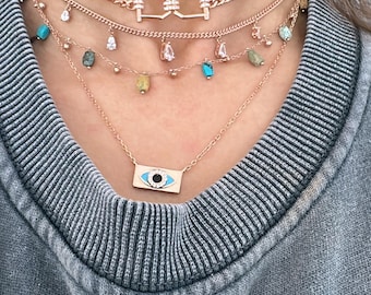 May include: A layered necklace set with a rose gold chain, a charm with a diamond-like design, and a pendant with a blue evil eye design. The necklace is worn on a person with a grey sweater.