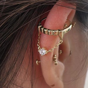 May include: Gold-toned ear cuff with a chain and a dangling clear gemstone. The cuff wraps around the ear's cartilage, connected by a delicate chain to a stud earring. Additional small gold studs are also visible.