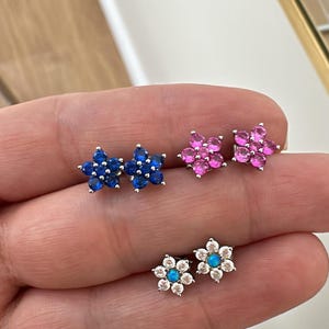May include: Three pairs of flower-shaped earrings with different colored gemstones. The first pair has blue gemstones, the second pair has pink gemstones, and the third pair has white gemstones with a blue center.
