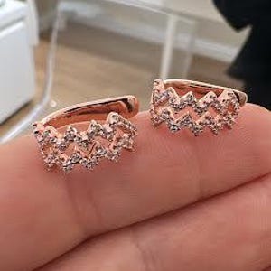 Op de afbeelding: Roségouden oorringen met een zigzagpatroon, versierd met kleine, glinsterende steentjes. De oorbellen worden op een hand getoond, waarbij hun fijne details en elegante stijl worden benadrukt. Een modieus accessoire.