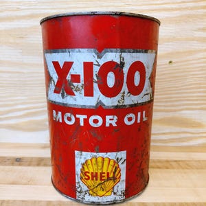 May include: Vintage red Shell X-100 Motor Oil can. The can features white and red lettering, with the words "X-100" prominently displayed. The Shell logo is visible at the bottom. The can shows signs of wear and tear.