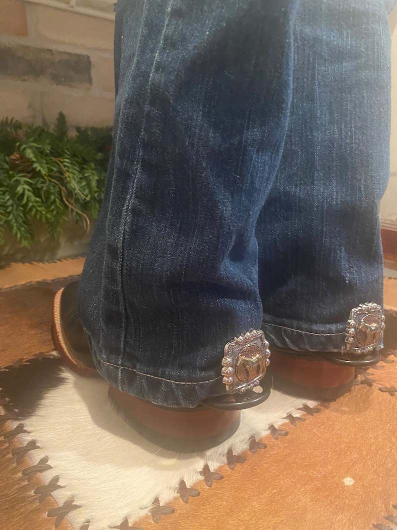 May include: A pair of blue jeans with silver and gold metal boot spurs attached to the bottom of the legs. The spurs are on a white and brown cowhide rug.