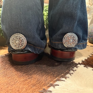 May include: Close-up of the lower portion of dark blue denim jeans with decorative silver-toned embellishments near the hem. The jeans are paired with brown leather boots with a dark red sole. The background features a cowhide rug.