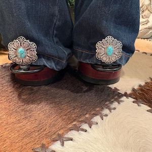 Puede incluir: Primer plano de vaqueros con clips decorativos de filigrana plateada con una piedra turquesa en el centro. Los clips est&aacute;n sujetos a la parte inferior de los vaqueros, apoyados en zapatos burdeos. El fondo incluye una alfombra de piel de vaca.