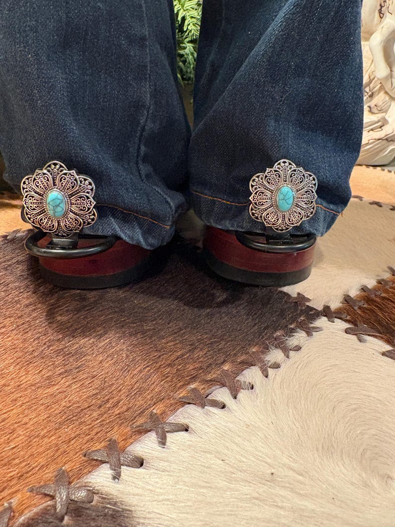 Puede incluir: Primer plano de vaqueros con clips decorativos de filigrana plateada con una piedra turquesa en el centro. Los clips est&aacute;n sujetos a la parte inferior de los vaqueros, apoyados en zapatos burdeos. El fondo incluye una alfombra de piel de vaca.