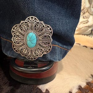 Puede incluir: Un clip de bota de metal plateado con una piedra turquesa ovalada en el centro. El clip tiene un dise&ntilde;o floral de filigrana y est&aacute; sujeto a una pernera de vaquero oscura. El clip de la bota est&aacute; sujeto a una bota marr&oacute;n y negra.