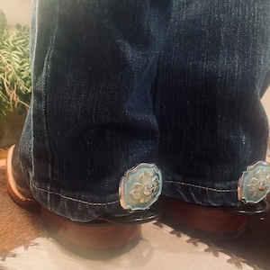 May include: A pair of blue jeans with silver and light blue metal boot straps. The straps have a decorative floral design.