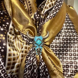 May include: A patterned scarf in shades of gold, brown, and white, secured with a turquoise and silver bull skull scarf slide. The scarf features geometric and paisley-like designs, creating a stylish accessory.