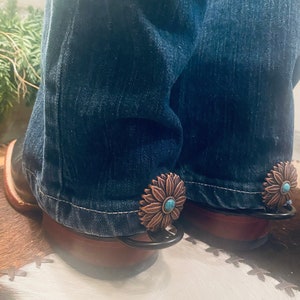 Op de afbeelding: Een paar blauwe jeans met koperen en turquoise bloemvormige sporen bevestigd aan de onderkant van de pijpen.