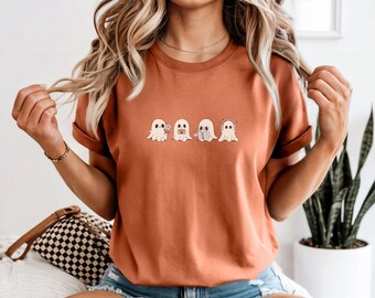 Op de afbeelding: Een roestkleurig T-shirt met vier cartoongeesten met verschillende uitdrukkingen en accessoires. De geesten zijn horizontaal gerangschikt. Het woord "Yam" staat op een witte rechthoek onderaan de afbeelding.