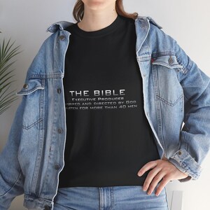 Puede incluir: Camiseta negra con el texto "THE BIBLE" en letras plateadas. Debajo, el texto dice "EXECUTIVE PRODUCER INSPIRED AND DIRECTED BY GOD WRITTEN FOR MORE THAN 40 MEN". La camiseta se usa con una chaqueta vaquera azul.