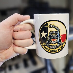 Może przedstawiać: Biały kubek ceramiczny z czarno-żółtym kolistym wzorem przedstawiającym zwiniętego węża z czerwoną i białą flagą Teksasu w tle. Pod wężem znajduje się napis "Don't Tread On Me".