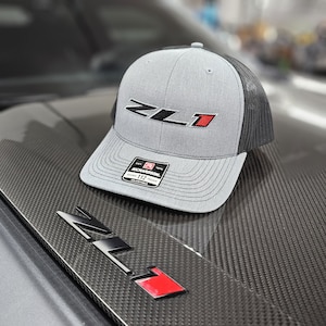 May include: A gray and black trucker hat with a red and black "ZL1" logo embroidered on the front. The hat is on a black carbon fiber surface.