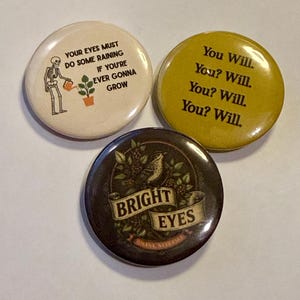 May include: Three circular pinback buttons. One cream button features a skeleton watering a plant with the text: YOUR EYES MUST DO SOME RAINING IF YOU'RE EVER GONNA GROW. One mustard yellow button reads: You Will. You? Will. You? Will. You? Will. One dark brown button shows a bird and foliage with the text: BRIGHT EYES OMAHA NEBRASKA.