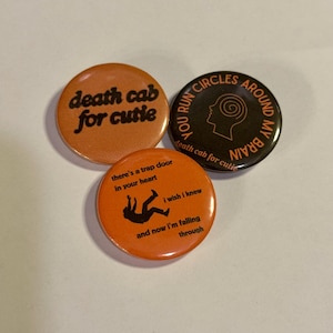 May include: Three circular pin-back buttons. One is orange with black text reading death cab for cutie. Another is black with orange text reading YOU RUN CIRCLES AROUND MY BRAIN and death cab for cutie. The third is orange with black text and a silhouette.