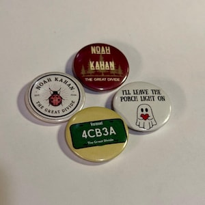 May include: Four circular pinback buttons featuring Noah Kahan designs. One is maroon with text, one is white with a ladybug, one is white with a ghost holding a heart and the text I'LL LEAVE THE PORCH LIGHT ON, and one is yellow with a Vermont license plate.