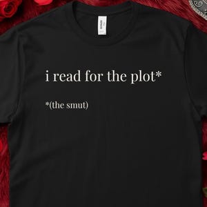 May include: Black t-shirt with the text "i read for the plot* *(the smut)" in white. The shirt has a crew neck and short sleeves. The shirt is made of a soft material.