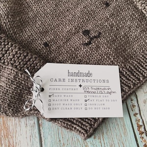 A brown baby sweater with a face embroidered on the front. A handmade care instruction label is tied to it's right sleeve.