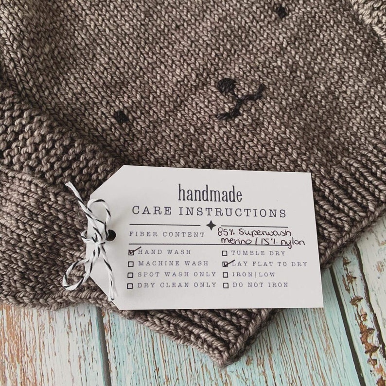 A brown baby sweater with a face embroidered on the front. A handmade care instruction label is tied to it's right sleeve.