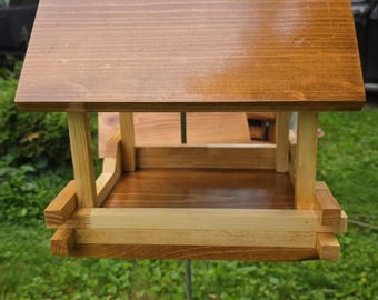 Può includere: Mangiatoia per uccelli in legno con tetto tinto di marrone e dettagli in legno naturale. La mangiatoia è sospesa da una catena e ha un vassoio per semi. Il design è semplice e rustico.