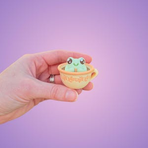 May include: A small, light green frog figurine with large eyes and a smiling mouth sits inside a yellow teacup. The teacup has a decorative leaf pattern around the rim and a small handle. The frog is the focal point of this whimsical, decorative item.