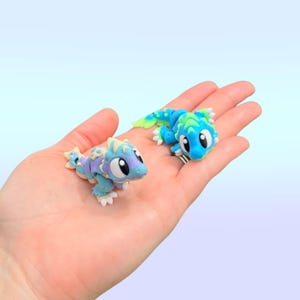 May include: Two small, colorful lizard figurines. One is light blue, purple, and cream, the other is blue and green. Both have large, expressive eyes and are posed on a light blue background.