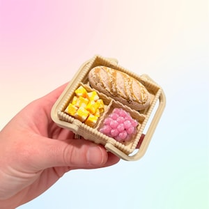 Puede incluir: Una cesta de picnic en miniatura con asa, que contiene un pan, cubos amarillos y esferas rosas. La cesta es de color beige y tiene un dise&ntilde;o tejido. Los alimentos parecen ser r&eacute;plicas a peque&ntilde;a escala.