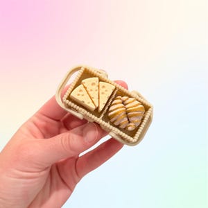 Puede incluir: Una peque&ntilde;a cesta de picnic beige con cu&ntilde;as de queso y cruasanes en miniatura. Los alimentos tienen texturas y colores detallados. El fondo es un suave degradado de rosa, amarillo y azul.