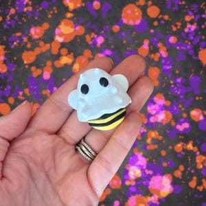 May include: A whimsical Halloween-themed figurine combining a ghost and a bee. The white ghost has black eyes and a cute expression, atop a yellow and black striped bee body. The background features a colorful splatter design.