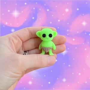 May include: A small, green alien figurine with large black eyes and a tan diaper. The alien has large ears and is standing upright. The background is a soft, colorful blend of pink, purple, and white.