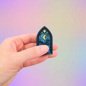 May include: A dark blue, arched-shaped pin featuring a stained-glass design with a yellow crescent moon, stars, and bats. The pin is held in a hand against a soft, pastel-colored background. The pin is a gothic-themed accessory.