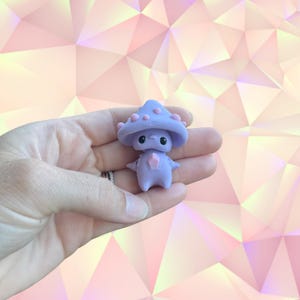 May include: A small, lavender mushroom creature figurine with a mushroom cap and a pink gem on its chest. The figurine has black eyes and is held in a hand. The background is a geometric pattern of pink and white.